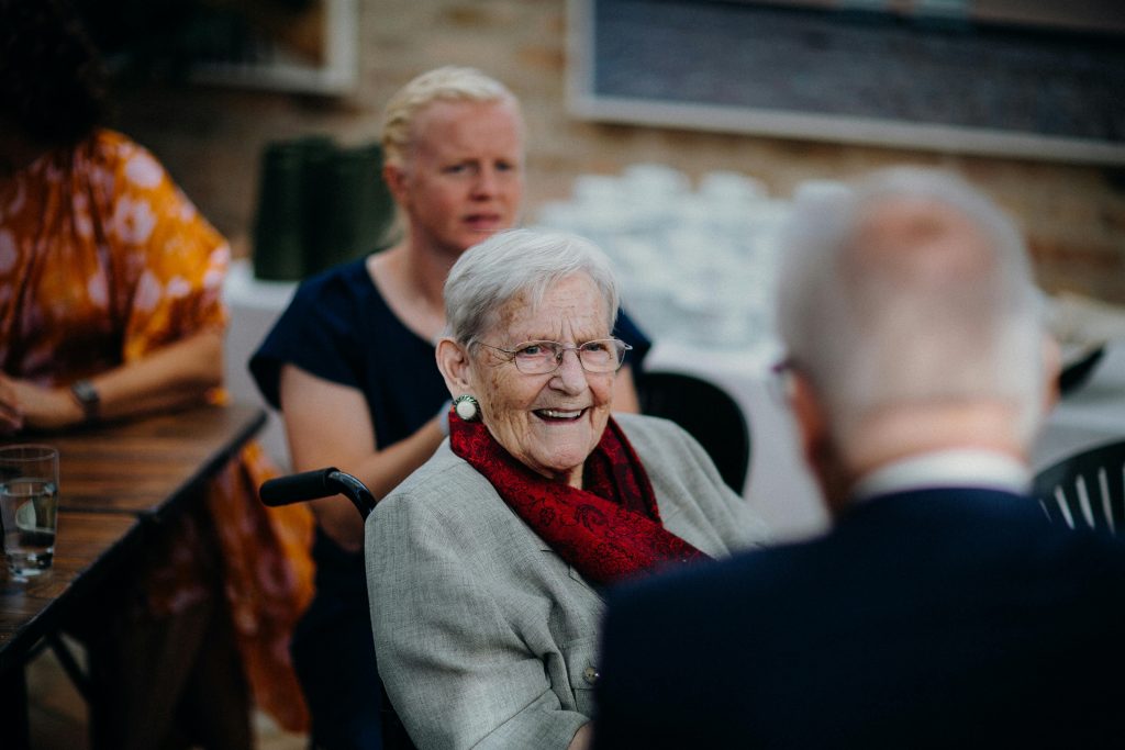 an elderly woman wearing a coat and a burgundy scarf smiling at someone out of frame